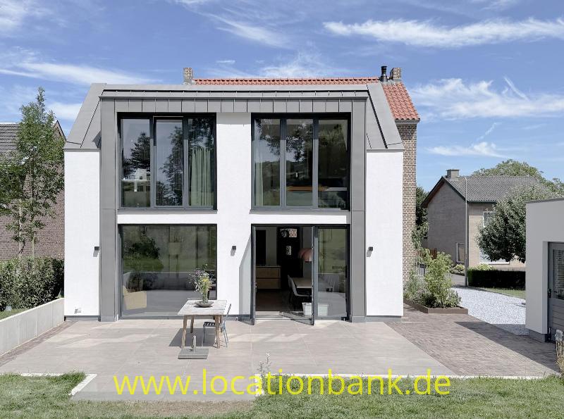 Hausfassade mit großen Fenstern, einem roten Ziegeldach und einer gepflasterten Terrasse.