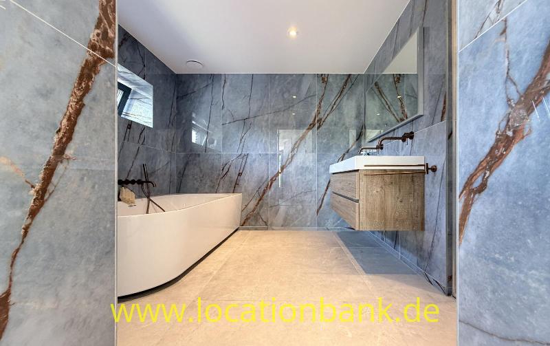 Badezimmer mit einer freistehenden weißen Badewanne, einem hölzernen Waschtischunterschrank und grauen Wandfliesen mit braunen Adern.