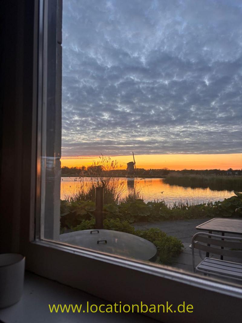 Ein von Schilf gesäumter See spiegelt den orangefarbenen Sonnenuntergang, gesehen durch ein Fenster.