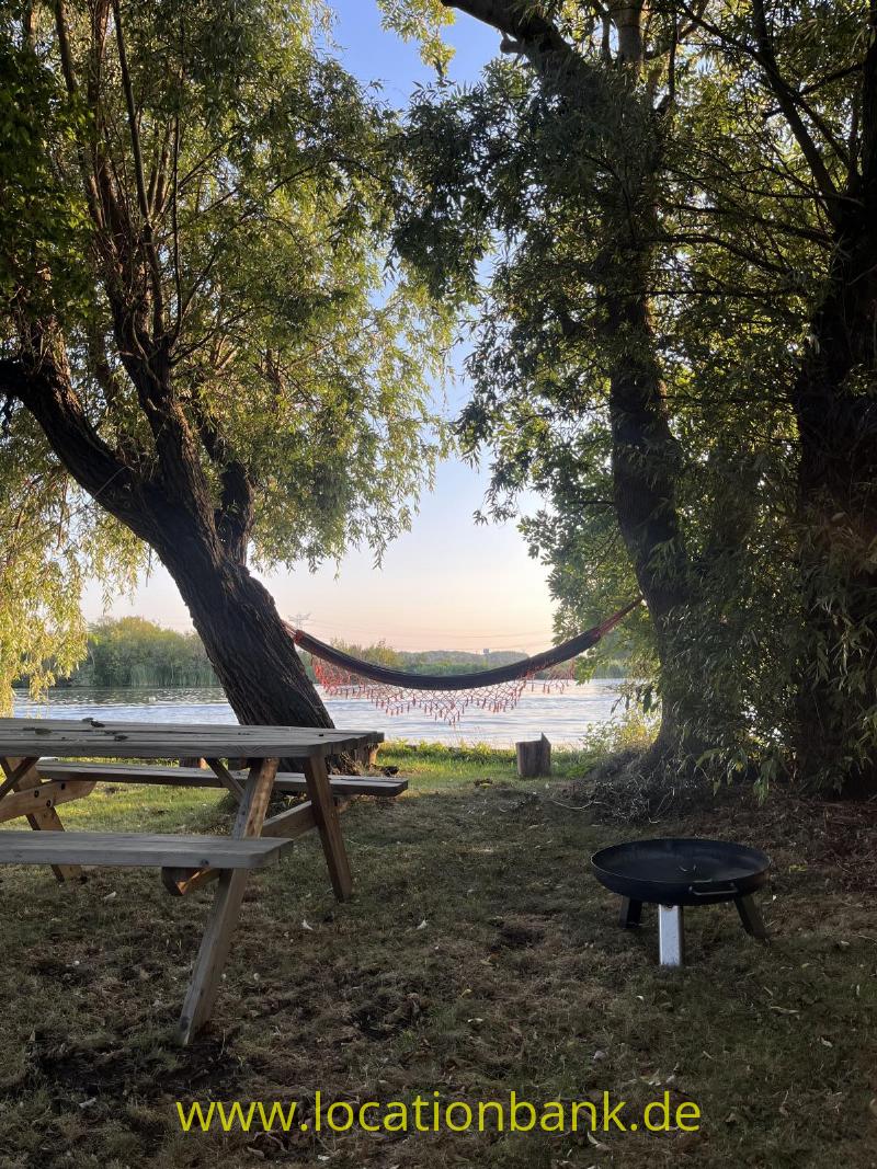 Ein hölzerner Picknicktisch steht unter Bäumen im Garten neben einer Hängematte am Wasser.