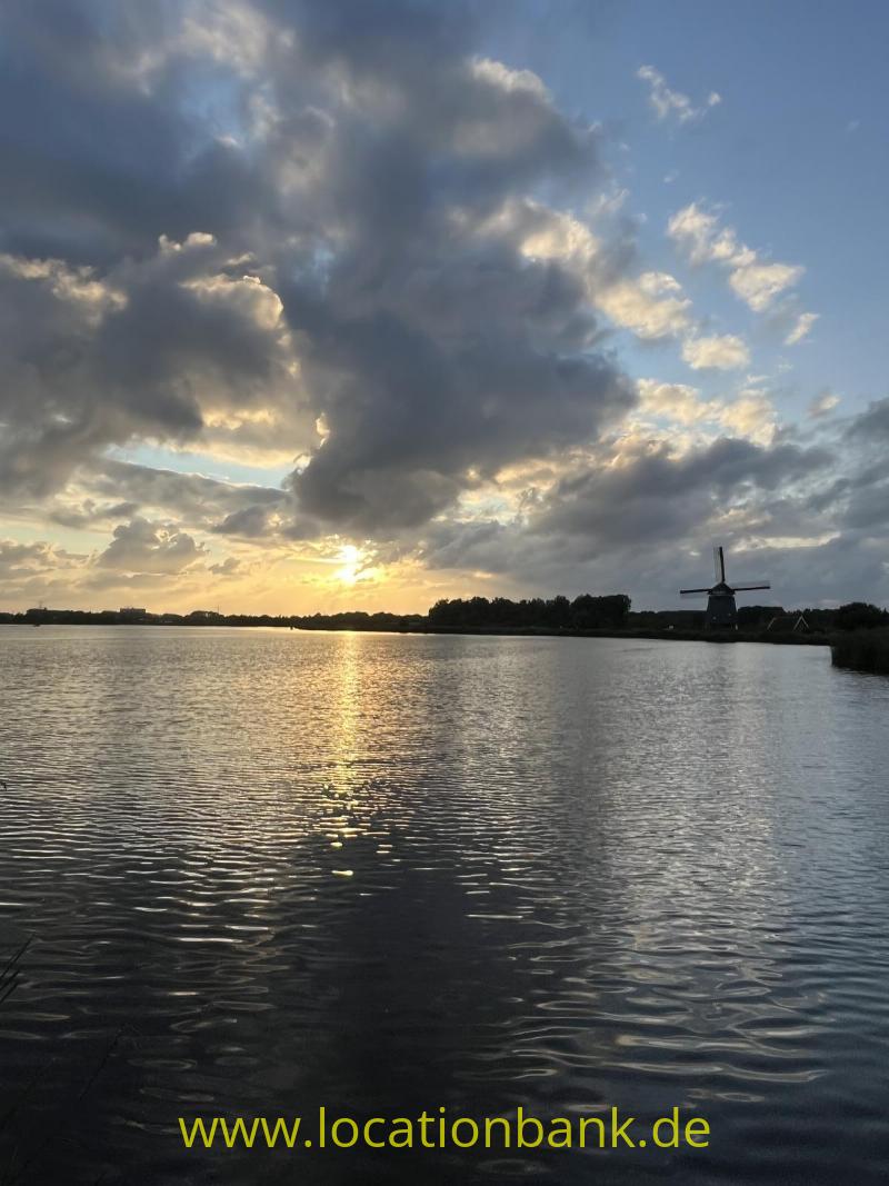 Eine Windmühle steht am Wasser, während die tief stehende Sonne durch dunkle Wolken scheint.