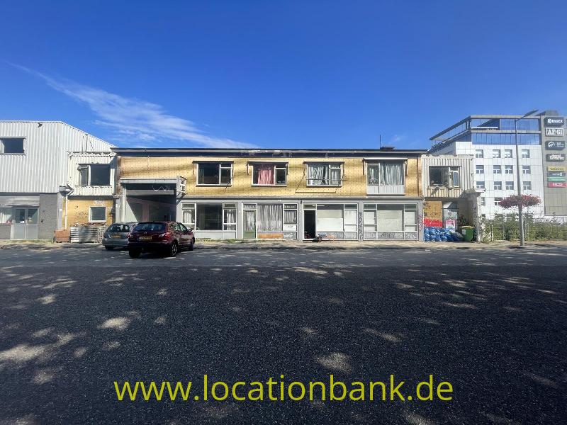 Zweigeschossiges Gebäude mit Balkonen und großen Fenstern, flankiert von Industriegebäuden und geparkten Autos.