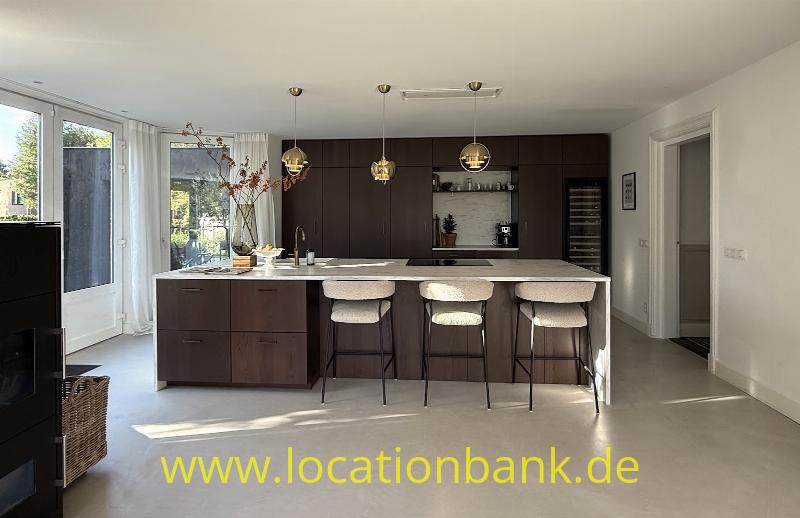 Küche mit einer großen Kochinsel, drei Barhockern, dunklen Schränken und drei Pendelleuchten neben hohen Fenstern.
