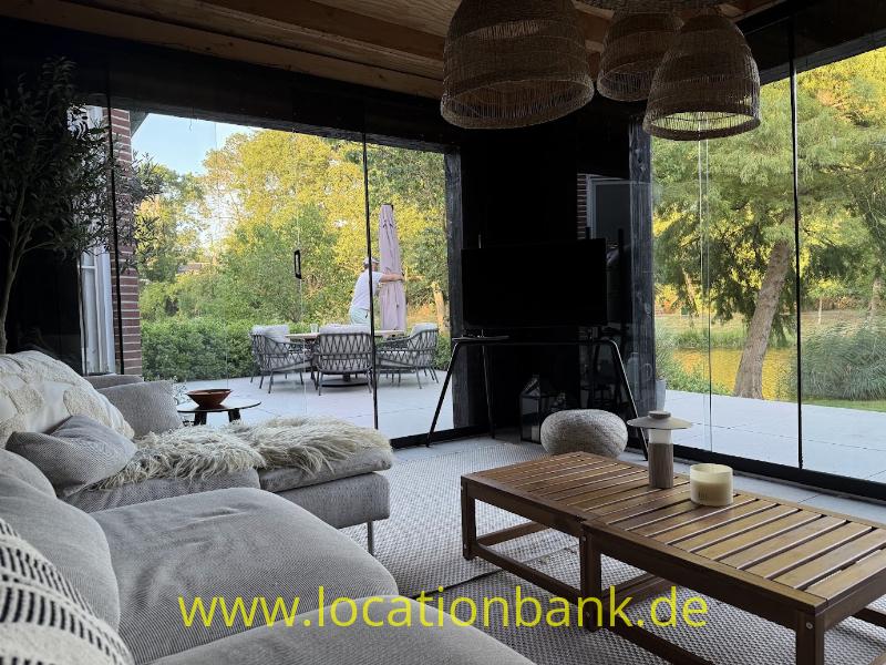 Wohnzimmer mit großem Ecksofa, hölzernem Couchtisch und bodentiefen Fenstern mit Blick auf einen Garten und eine Terrasse.