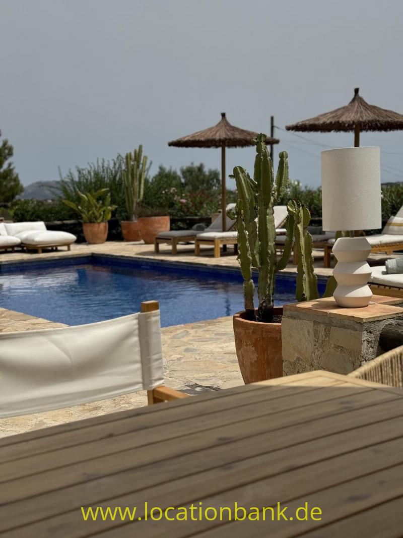 Swimmingpool mit Liegen, Sonnenschirmen, Kakteen, einer Außenleuchte und einer Holzterrasse im Vordergrund.
