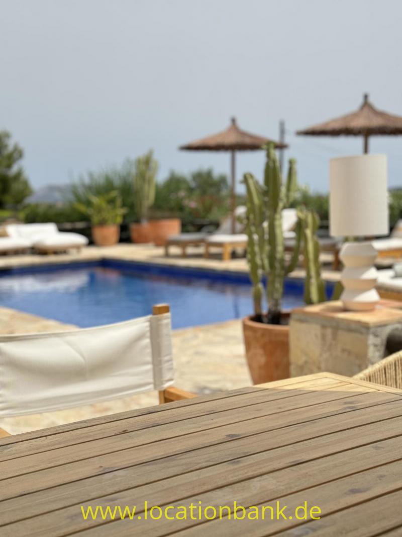 Holztisch im Vordergrund mit einem Swimmingpool, Sonnenschirmen und Topfpflanzen im Hintergrund.