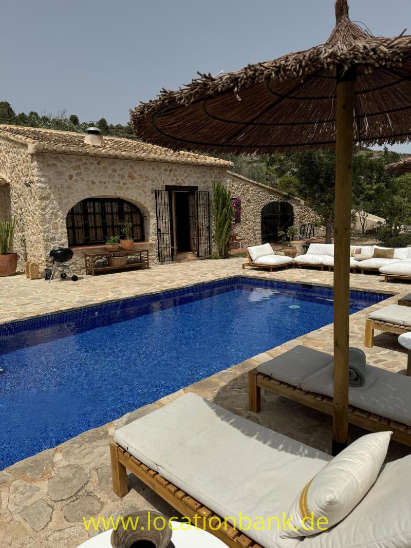 Blauer Swimmingpool in Spanien neben einem Steingebäude mit strohgedeckten Sonnenschirmen und Sonnenliegen.