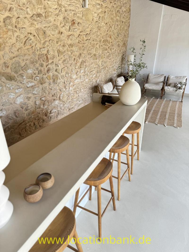 Lange beigefarbene Theke mit Holzstühlen, einer großen Vase und einer Natursteinwand.