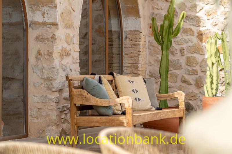 Ein Holzsessel mit Kissen steht vor einer Steinmauer neben hohen Kakteen.