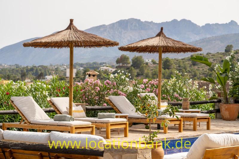 Sonnenliegen und Strohs­­chirme stehen zwischen blühenden Pflanzen neben einem kleinen Pool, im Hintergrund sind Berge zu sehen.