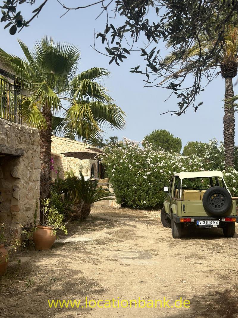 Ein grüner Geländewagen mit Reserverad steht auf einem unbefestigten Innenhof neben einem Steingebäude und Palmen.