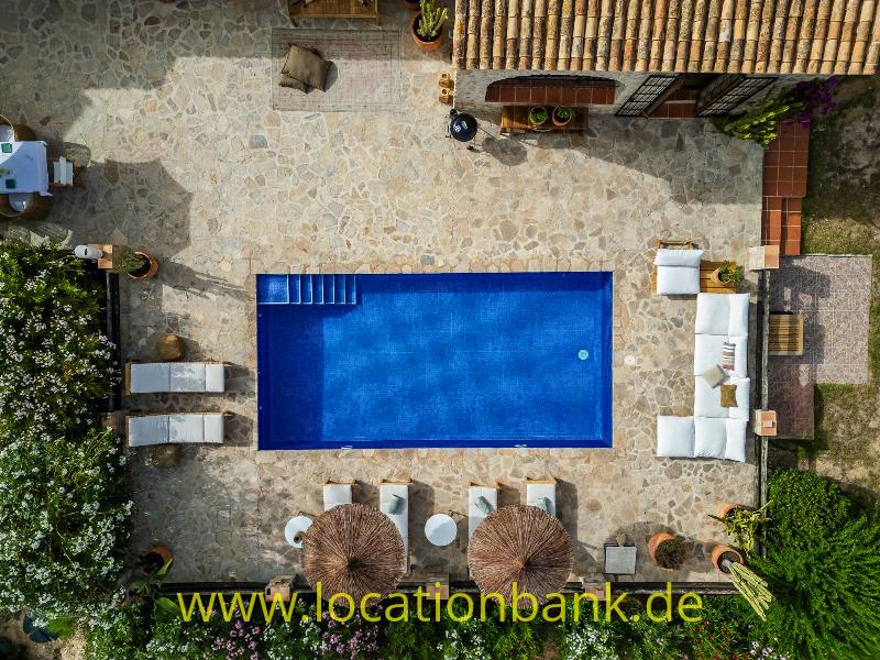 Rechteckiger blauer Swimmingpool in einem mit Naturstein gepflasterten Innenhof, umgeben von Liegestühlen, Sonnenschirmen und weißen Sitzmöbeln.