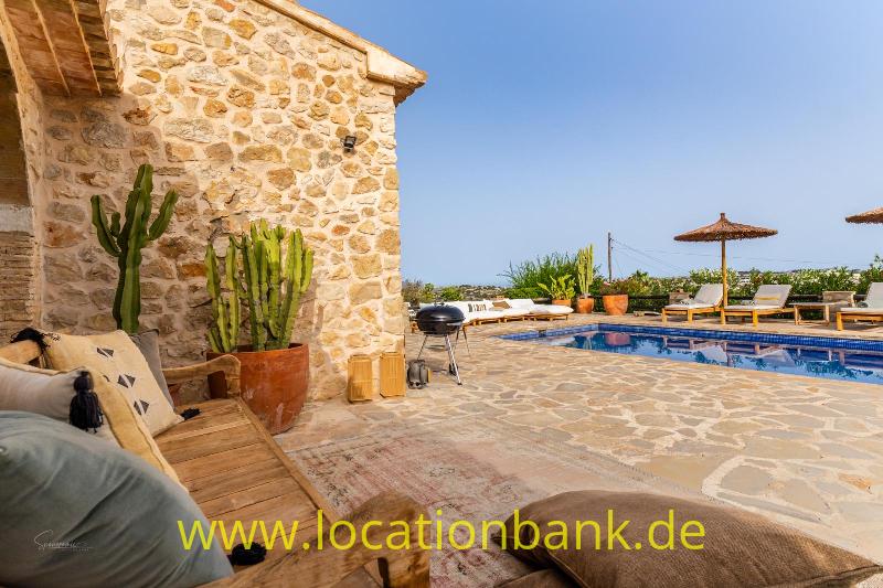 Steinterrasse neben einem Steingebäude mit Swimmingpool, Liegestühlen, Sonnenschirmen und Topfpflanzen.