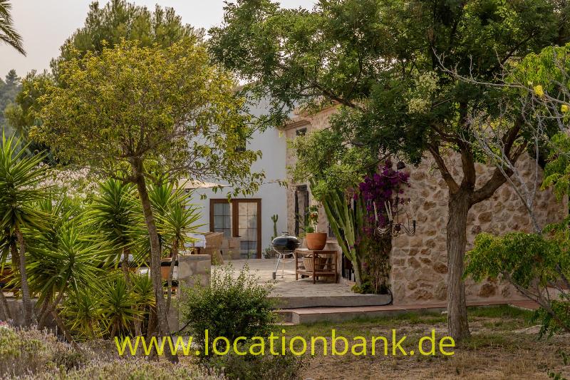 Garten mit Bäumen, Yuccas, blühenden Pflanzen und einer steinernen Gebäudefassade.