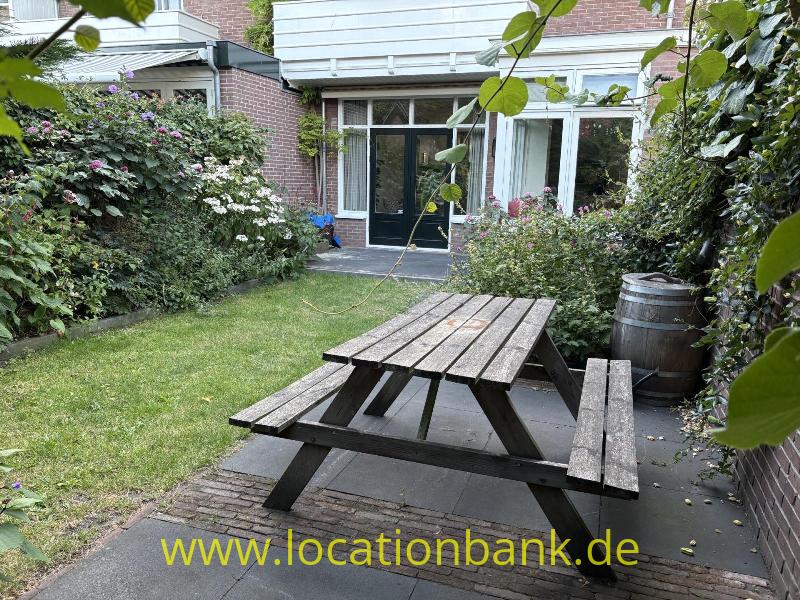 Ein hölzerner Picknicktisch steht auf einer gepflasterten Terrasse in einem grünen Innenhof neben einem Ziegelgebäude.