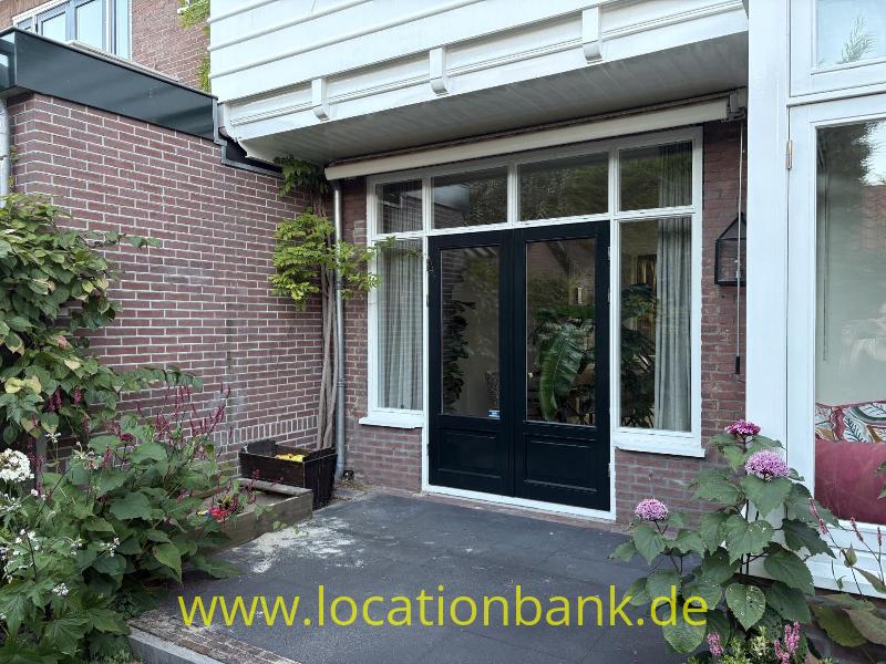Schwarze zweiflügelige Tür in einer Backsteinfassade, umgeben von weißen Fensterrahmen und blühenden Hortensien.