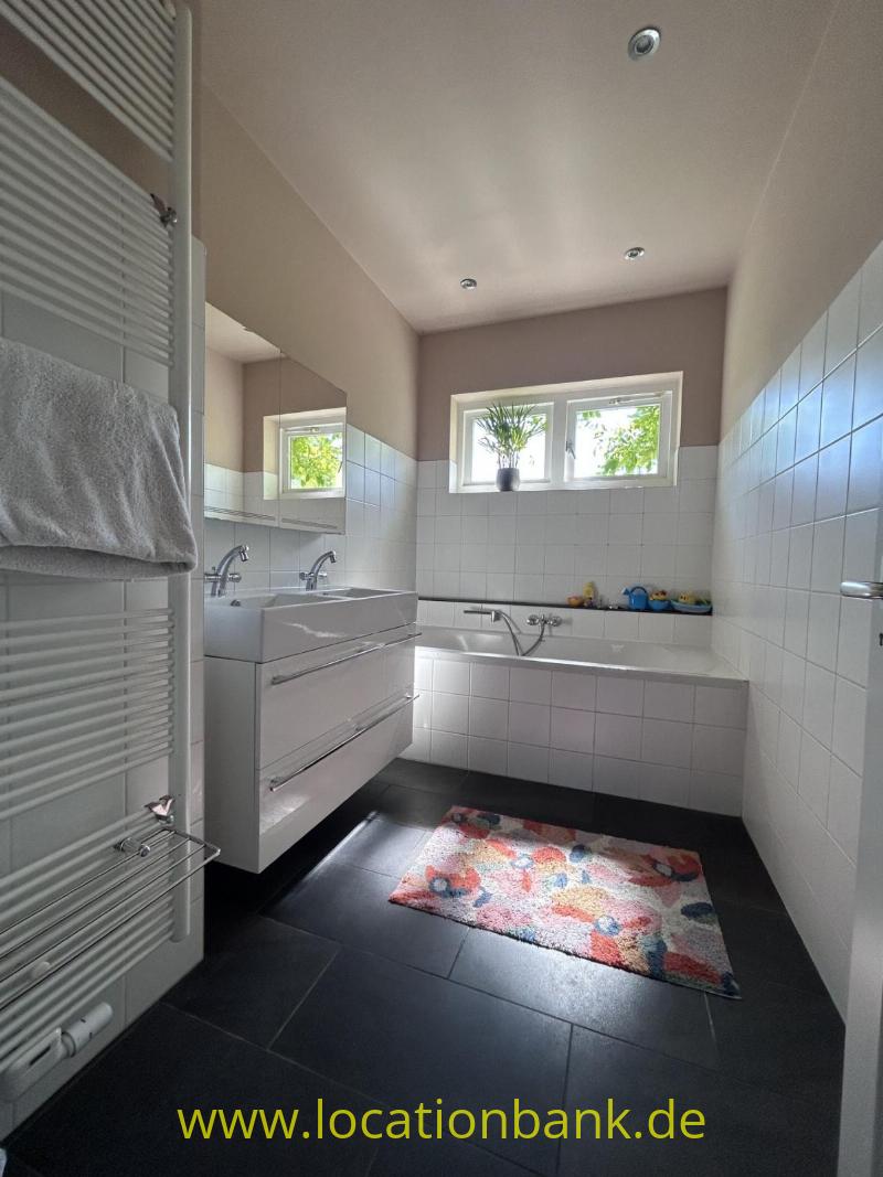 Badezimmer mit weiße Wandfliesen, eine Badewanne, einen Waschtischunterschrank, ein Fenster und einen bunten Teppich.