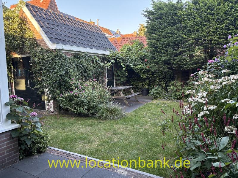 Garten mit einem hölzernen Picknicktisch, einem gepflasterten Weg, blühenden Pflanzen und einem Ziegeldach im Hintergrund.
