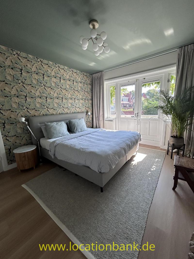 Schlafzimmer mit einem bezogenen Bett, botanischer Tapete, hohen Fenstern, Vorhängen und einer großen Zimmerpflanze.
