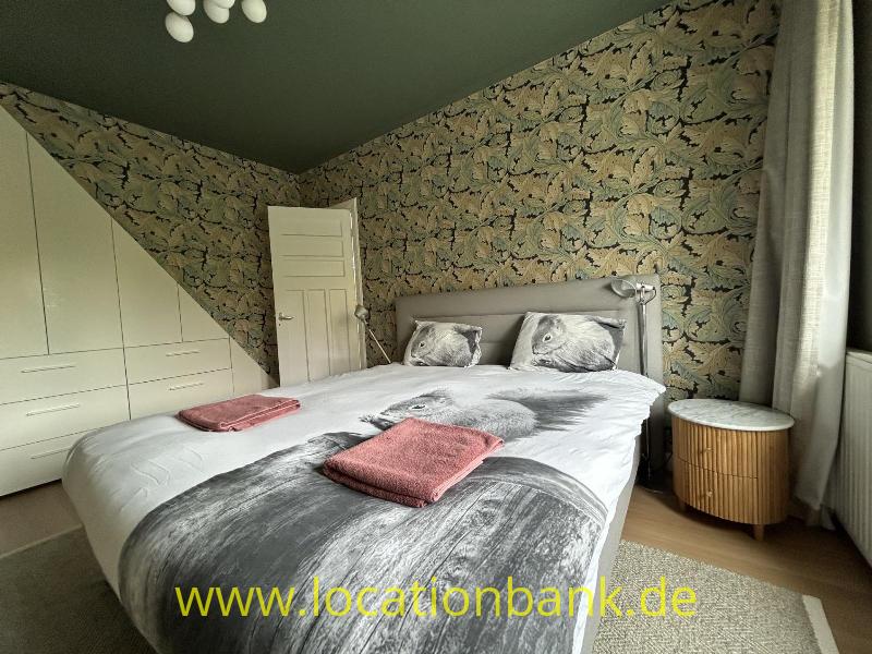 Schlafzimmer mit einem Doppelbett, botanischer Tapete, weißen Schranktüren und einem runden hölzernen Nachttisch.