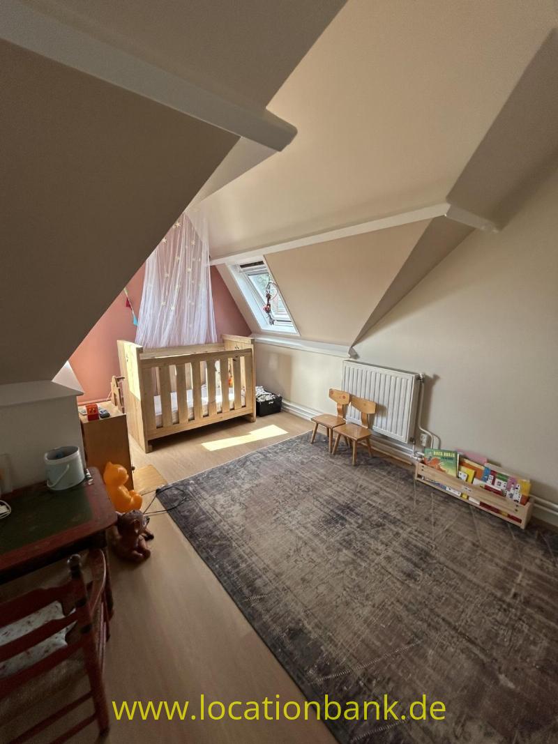 Ein hölzernes Kinderbett steht unter einer schrägen Decke neben einem Dachfenster, einem Teppich, einem Heizkörper und einem Spielzeugregal.