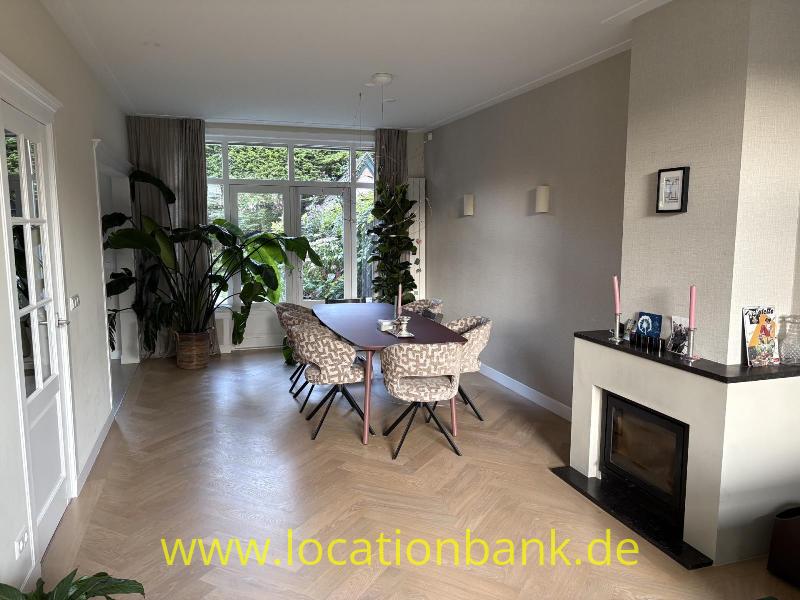 Esszimmer mit einem ovalen Tisch, mehreren Stühlen, großen Fenstern, Zimmerpflanzen und einem Kamin.