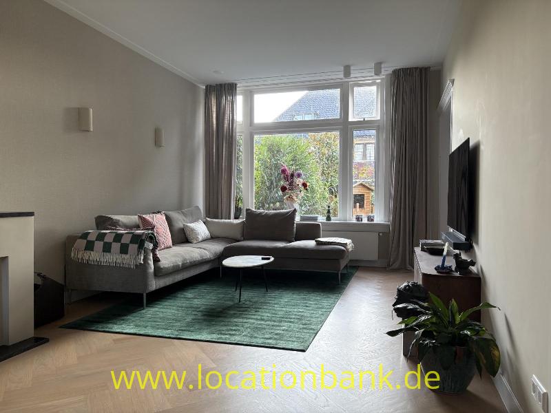 Wohnzimmer mit einem grauen Ecksofa, großen Fenstern, einem grünen Teppich und einem an der Wand montierten Fernseher.