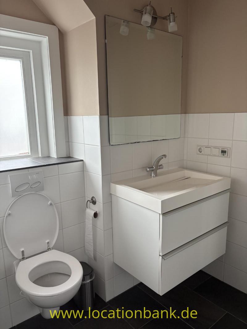 Badezimmer mit einem weißen, wandmontierten Waschtischunterschrank, Spiegel, Toilette und Fenster an gefliesten Wänden.