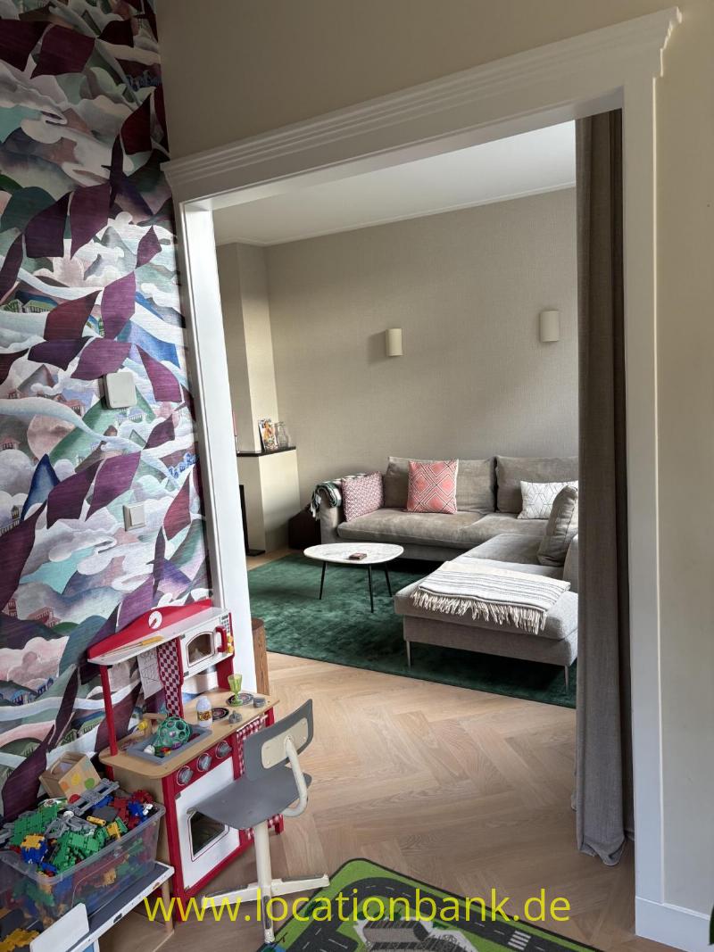 Durch eine Türöffnung ist ein Wohnzimmer mit grauem Ecksofa und grünem Teppich sichtbar.