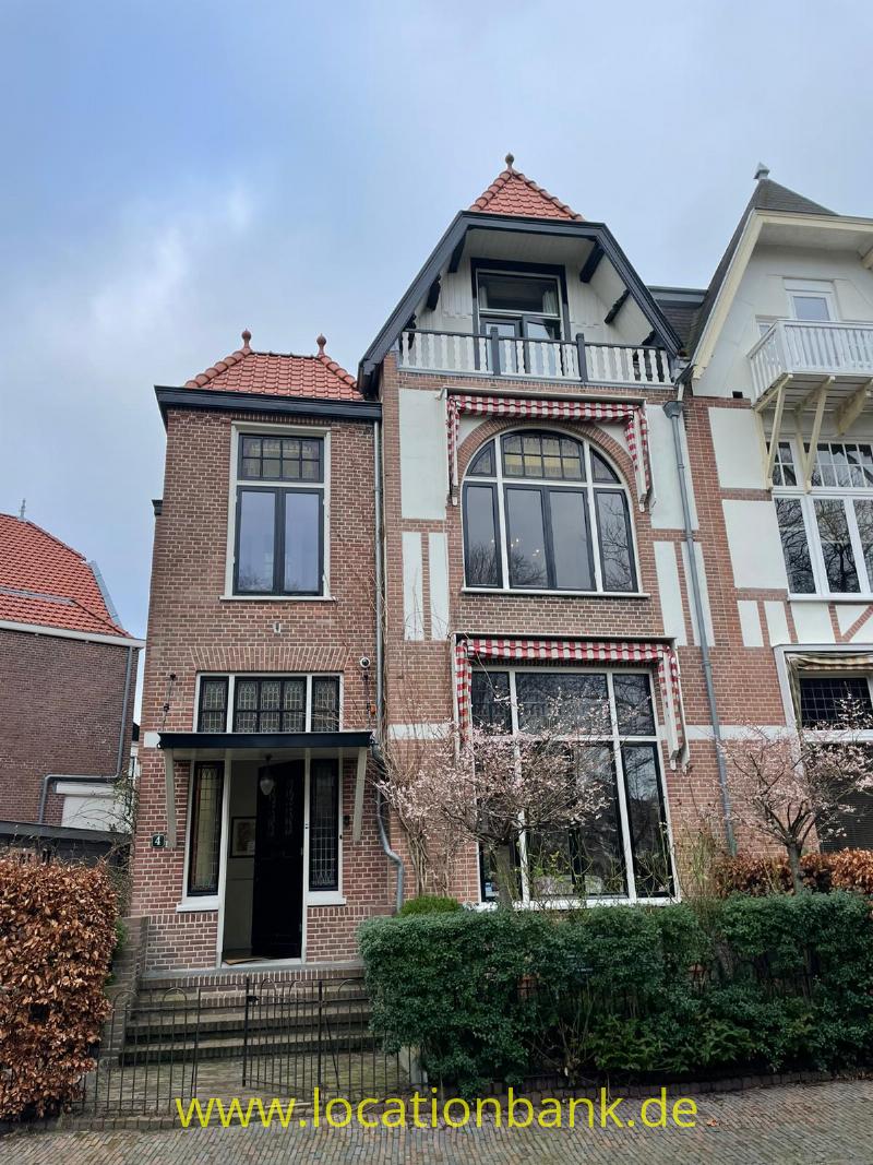 Backsteinhaus mit roten Ziegeldächern, weißen Balkonen und großen Rundbogenfenstern hinter einer Hecke.