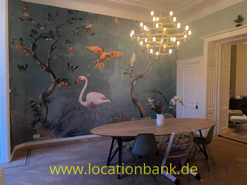 Raum mit einem Wandgemälde von Flamingos, Vögeln und blühenden Zweigen, einem ovalen Tisch und einem Kronleuchter.