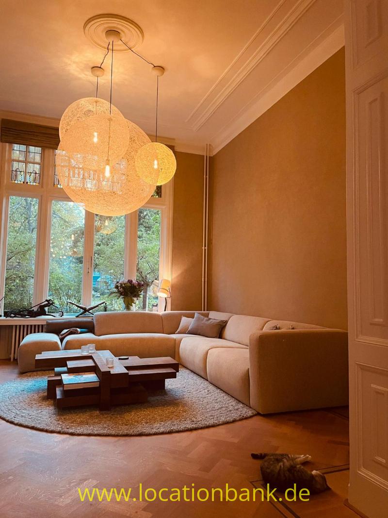Wohnzimmer mit einem geschwungenen beigen Ecksofa, drei hängenden Kugelleuchten und hohen Fenstern.