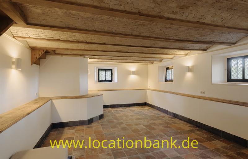 Geräumiger Raum mit sichtbaren Holzbalken, einem Boden aus Terrakottafliesen, kleinen Bleiglasfenstern und Holzbänken entlang der Wände.
