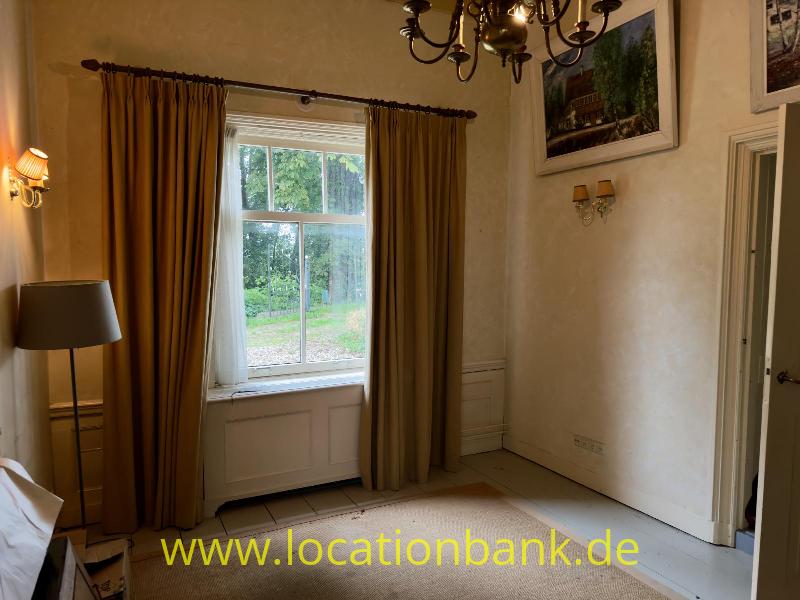 Zimmer mit hohen Fenstern, langen Vorhängen, einem Kronleuchter, Wandleuchten und gerahmten Gemälden.