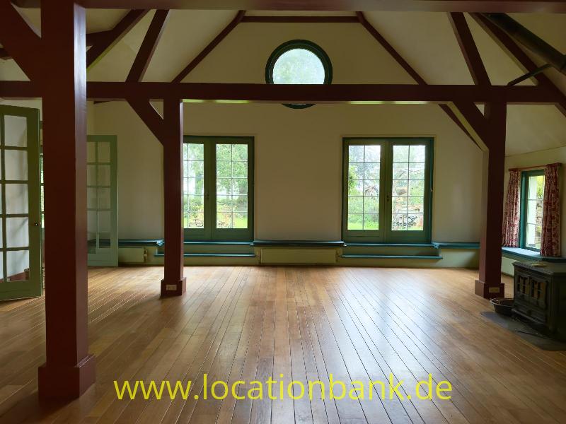 Geräumiger Raum mit sichtbaren dunklen Holzbalken, einem Holzboden, großen Fenstern und einem runden Fenster im oberen Wandbereich.