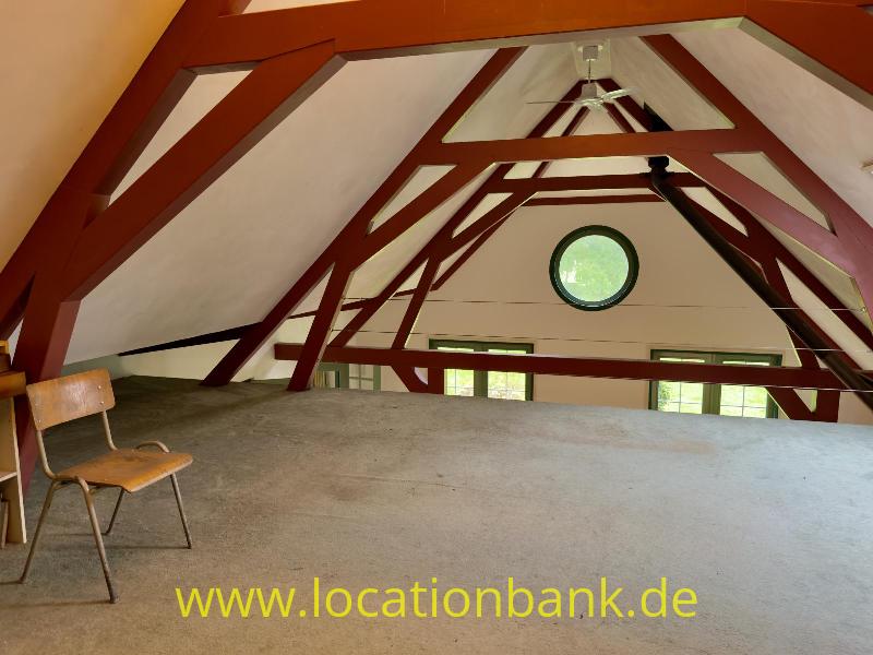 Ein Dachboden mit sichtbaren dunkelroten Dachbalken, einem runden Fenster und einem einzelnen Stuhl auf dem Boden.