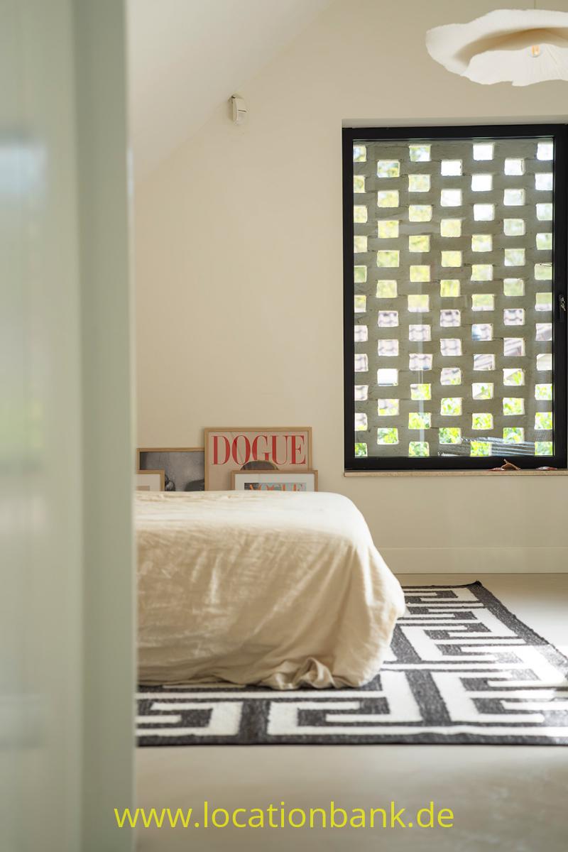 Ein Bett mit weißer Bettwäsche steht auf einem schwarz-weißen Teppich vor einem schwarz gerahmten Fenster mit einem Raster aus Quadraten.
