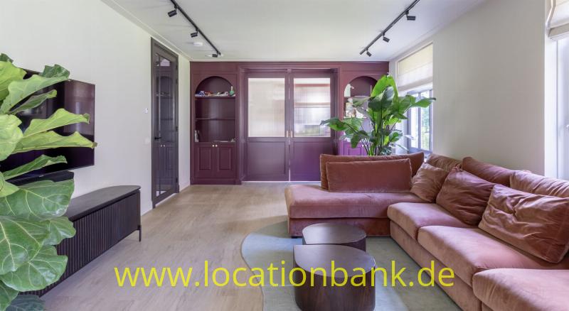 Wohnzimmer mit einem rosafarbenen Ecksofa, runden Holzcouchtischen, großen Pflanzen und dunklen Holzschränken.