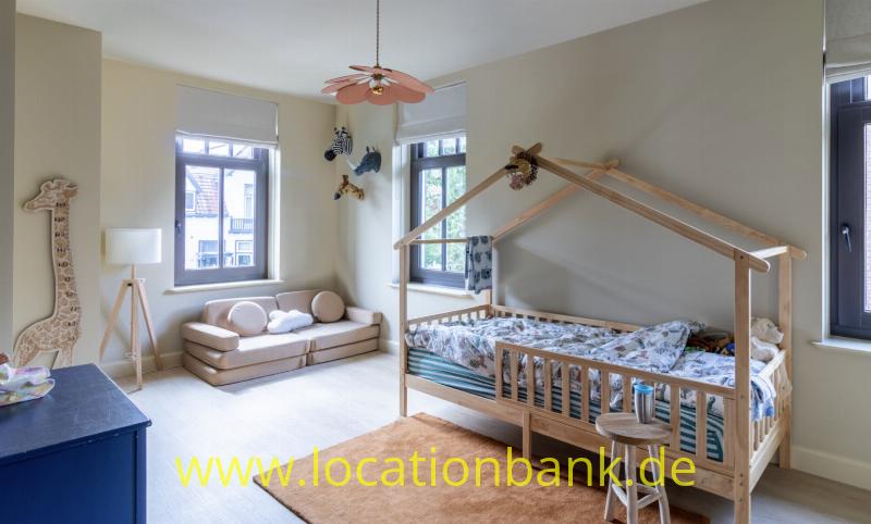 Holzbett mit hausförmigem Rahmen, blauer Bettwäsche und einem Hocker in einem hellen Zimmer.