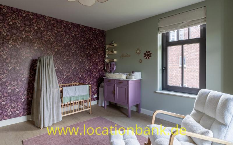 Zimmer mit dunkelvioletter Blumenmustertapete, einem Holz-Kinderbett, einer Wickelkommode, einem Sessel und einem großen Fenster.