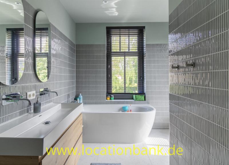 Freistehende weiße Badewanne in einem Badezimmer mit grauen Wandfliesen, zwei ovalen Spiegeln und Fenstern mit Jalousien.