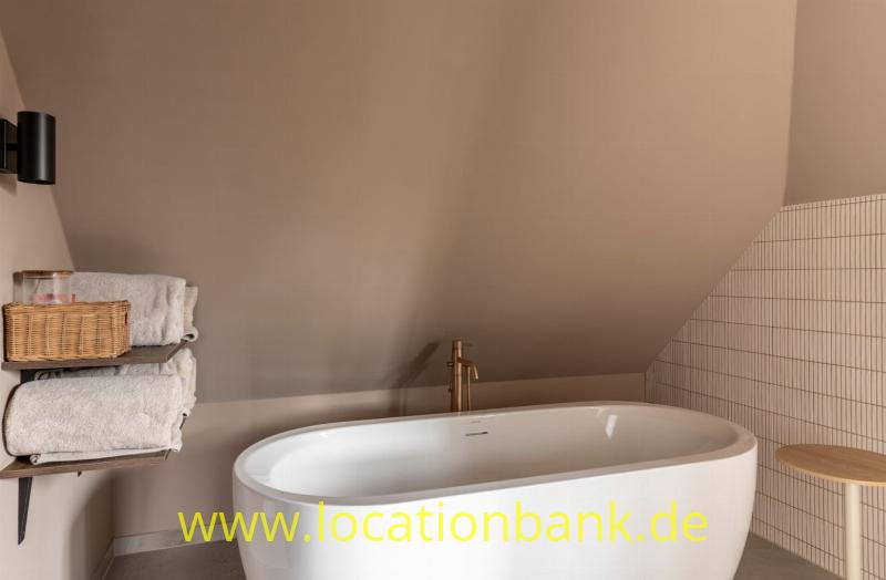 Freistehende weiße Badewanne unter einer Dachschräge, daneben gerollte Handtücher und ein Holzhocker.
