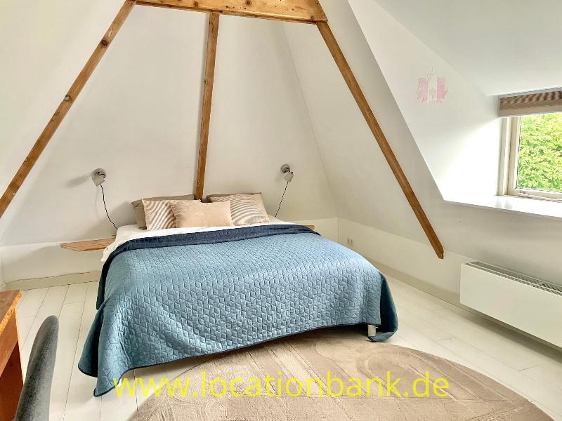 Ein Doppelbett mit einer blauen Tagesdecke steht unter Holzbalken in einem Raum mit Dachschräge und Fenster.