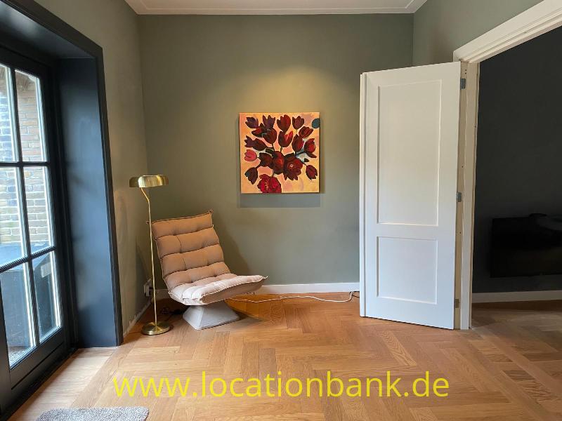 Sitzecke mit einem Loungesessel, einer Stehlampe, einem gerahmten Blumenbild und einem Fischgrätboden neben einer offenen weißen Tür.