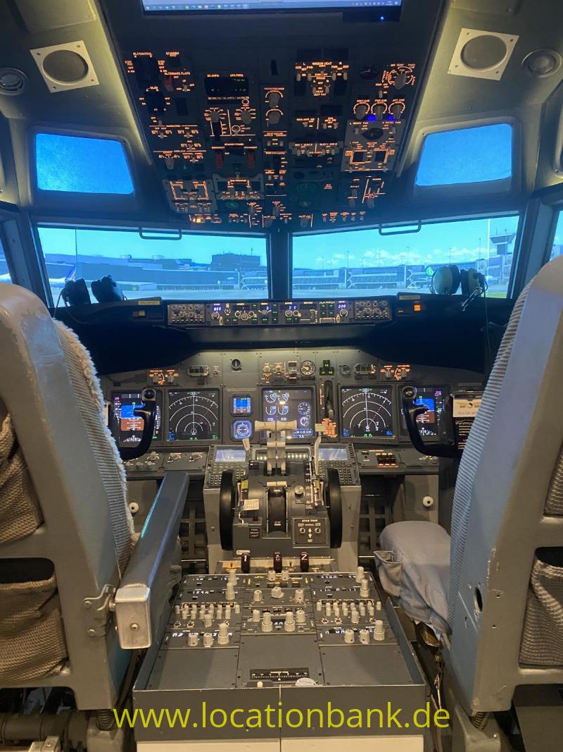Das Cockpit zeigt Bedienfelder, Steuerhörner und ein beleuchtetes Overhead-Panel; durch die Fenster ist Flughafen infrastruktur sichtbar.