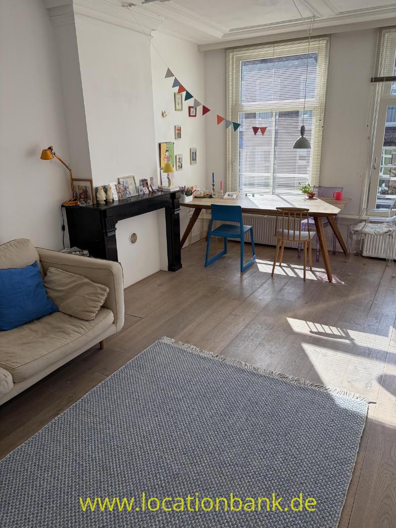 Wohnzimmer mit einem großen Fenster mit Jalousien, einem Schreibtisch, zwei Stühlen, einem Sofa und einer schwarzen Kaminverkleidung.