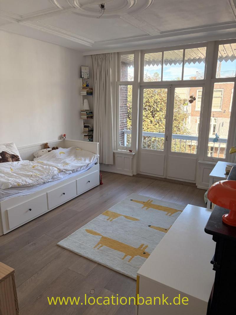 Schlafzimmer mit einem weißen Bett, hohen Fenstern, einem Teppich mit gelben Giraffen und einem Bücherregal.