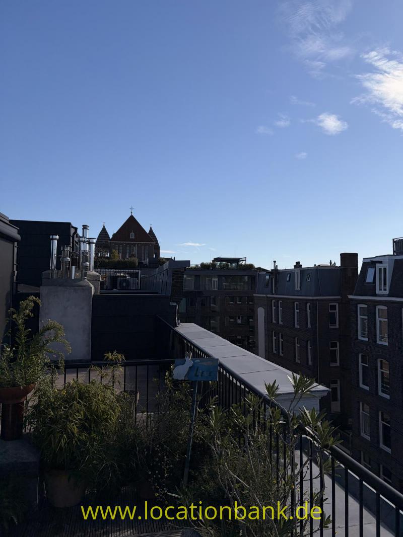 Dachterrasse mit Pflanzkübeln und Metallgeländer, umgeben von Backsteingebäuden und einem spitzen Turm im Hintergrund.