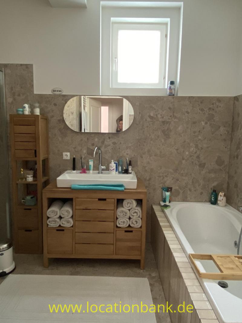 Badezimmer mit einem Waschtischmöbel aus Holz, einem runden Spiegel, einer Badewanne, beige gefliesten Wänden und einem Fenster.
