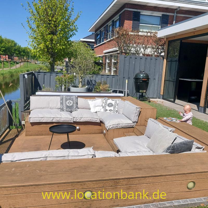 Holzterrasse am wasser mit einer Sitzgruppe, Pflanzgefäßen, einem Grill und Blick auf einen schmalen Kanal neben einem Haus.